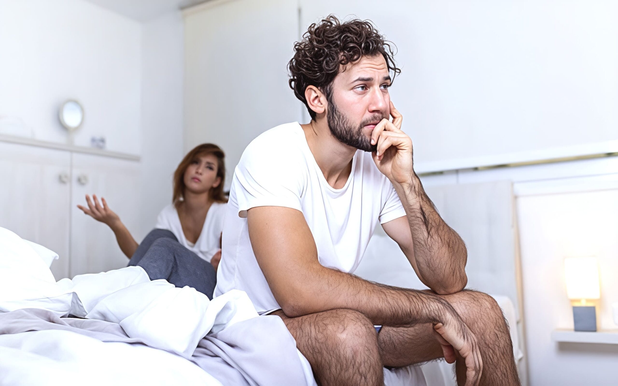 A man sitting on a bed with feet on the ground. His hand is holding his head in a thoughtful way. A woman is in the background addressing the man.
